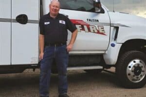 A man in a work uniform stands in front of a Falcon Fire truck equipped with a large crane arm.