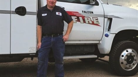 A man in a work uniform stands in front of a Falcon Fire truck equipped with a large crane arm.