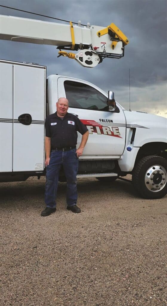 A man in a work uniform stands in front of a Falcon Fire truck equipped with a large crane arm.