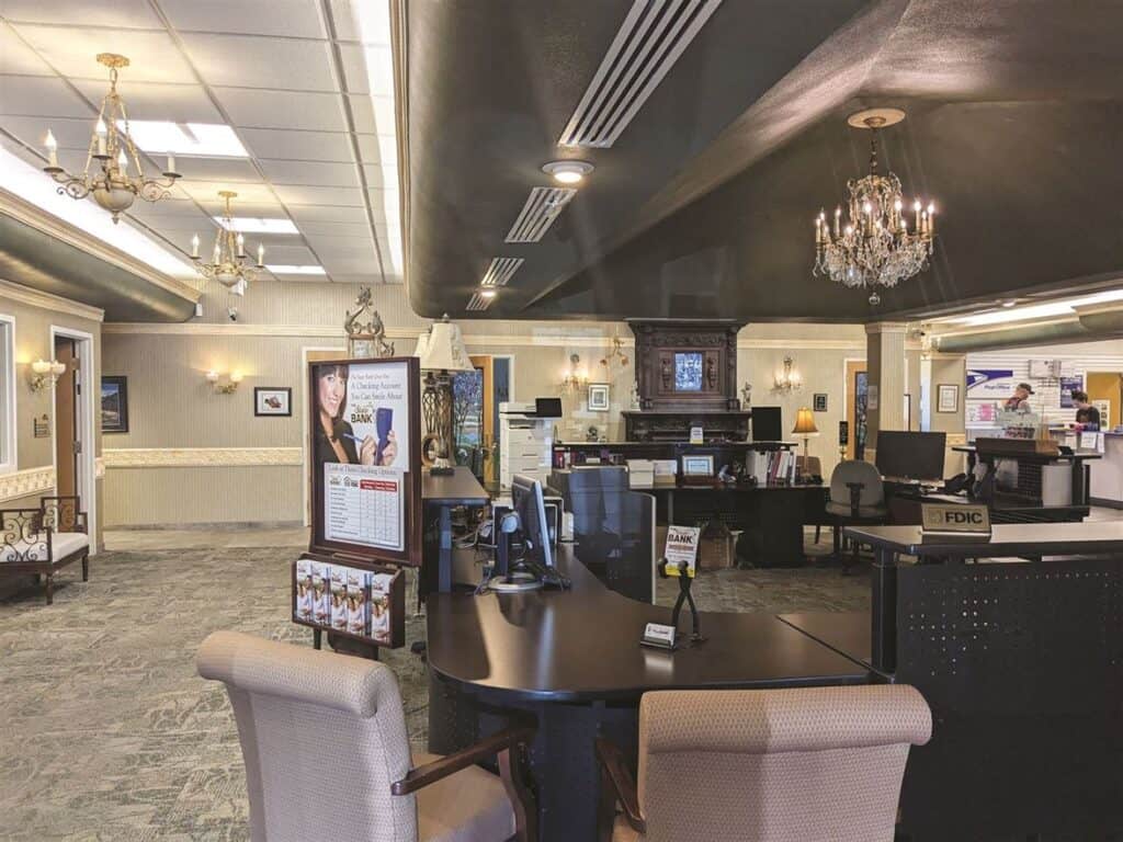 Interior of a bank featuring desks, chairs, posters, and chandeliers. A customer service counter is visible in the background. The decor is traditional with light-colored walls and carpeted floors.