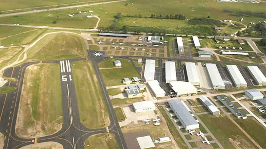 Aerial view of a small airfield with multiple runways, hangars, and parked light aircraft amidst surrounding green fields.