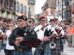 A group of musicians in traditional attire playing bagpipes marches through a busy, European-style street lined with buildings and onlookers.