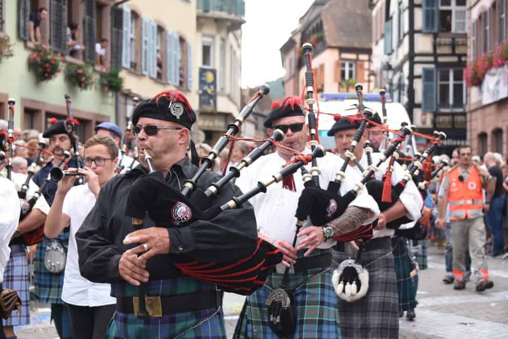 A group of musicians in traditional attire playing bagpipes marches through a busy, European-style street lined with buildings and onlookers.