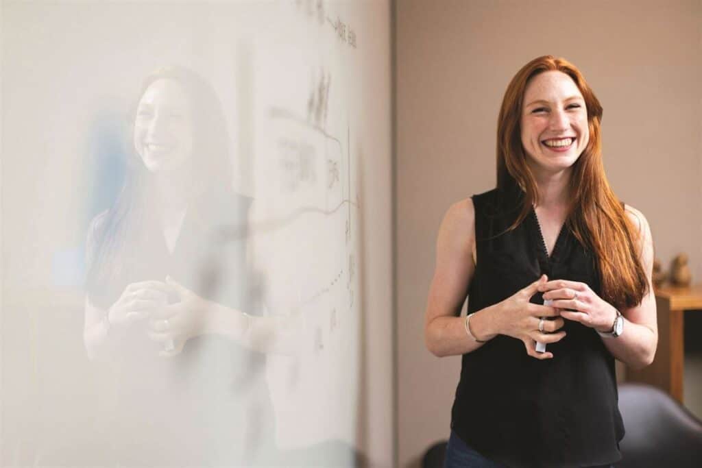 A woman with long red hair is standing and smiling near a whiteboard. She is wearing a black sleeveless top and has her hands clasped. Her reflection is visible on the whiteboard.