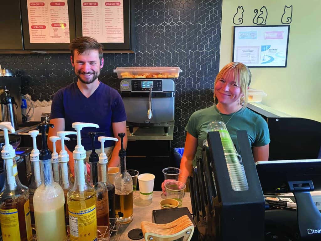 Two baristas, a man and a woman, standing behind a coffee shop counter with various syrup bottles and equipment.