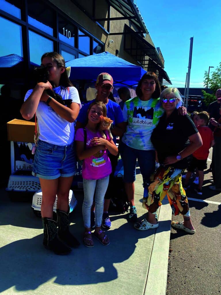 A group of five people and a child stand outside a building, smiling. One woman holds a small brown dog. A booth with a blue canopy is visible in the background.