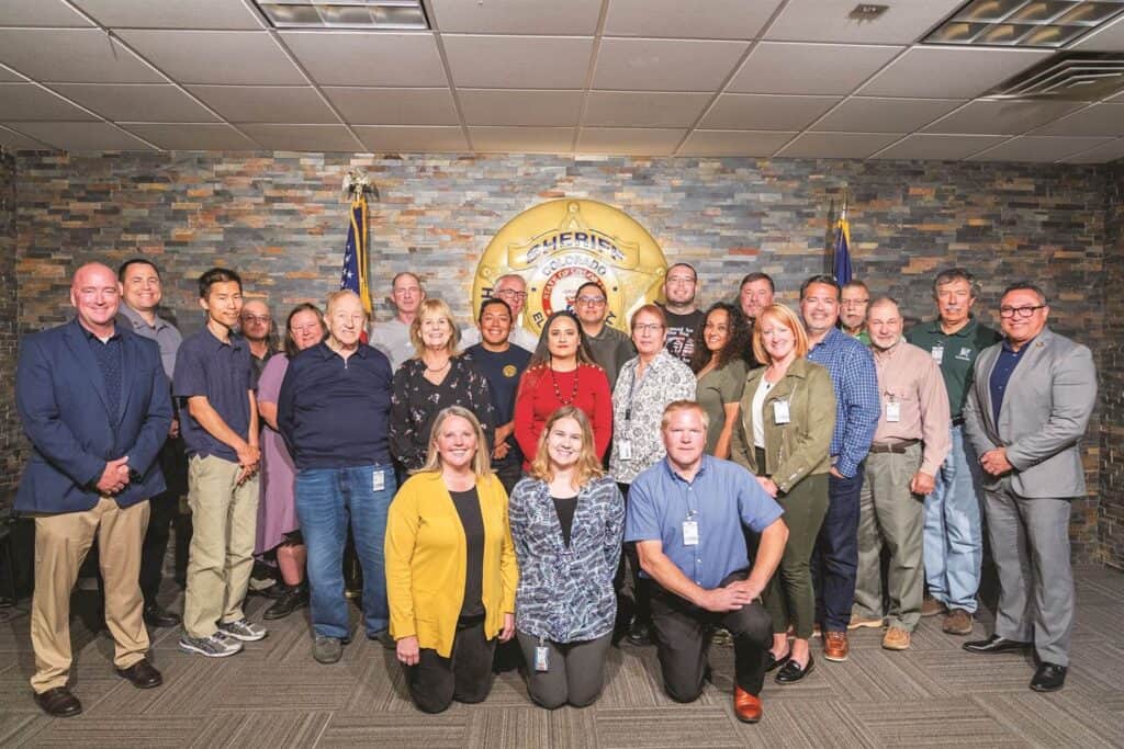 A group of 20 people stands in a room with a sheriff's badge emblem on the wall, posing for a group photo. They are dressed in casual and business attire.