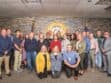 A group of 20 people stands in a room with a sheriff's badge emblem on the wall, posing for a group photo. They are dressed in casual and business attire.