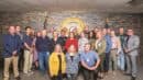 A group of 20 people stands in a room with a sheriff's badge emblem on the wall, posing for a group photo. They are dressed in casual and business attire.