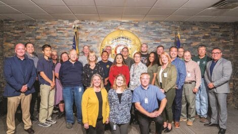 A group of 20 people stands in a room with a sheriff's badge emblem on the wall, posing for a group photo. They are dressed in casual and business attire.