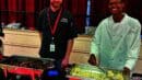 Two people in chef uniforms standing behind buffet trays with food. One smiles while serving, and the other looks ahead. Red curtain background.