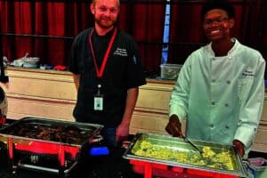 Two people in chef uniforms standing behind buffet trays with food. One smiles while serving, and the other looks ahead. Red curtain background.