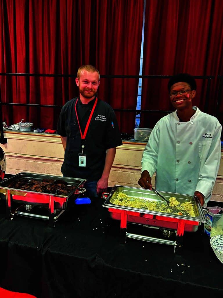 Two people in chef uniforms standing behind buffet trays with food. One smiles while serving, and the other looks ahead. Red curtain background.