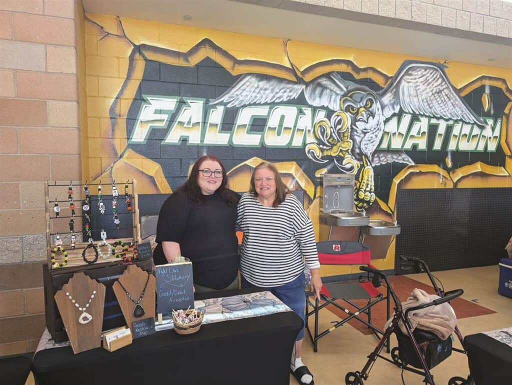 Two women stand behind a jewelry display table in front of a "Falcon Nation" mural.