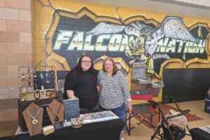 Two women stand behind a jewelry display table in front of a "Falcon Nation" mural.