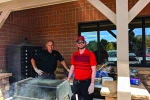 Two people standing by a grill outdoors, one wearing a red shirt and cap, both smiling. Brick building and tables with food items in the background.