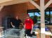 Two people standing by a grill outdoors, one wearing a red shirt and cap, both smiling. Brick building and tables with food items in the background.