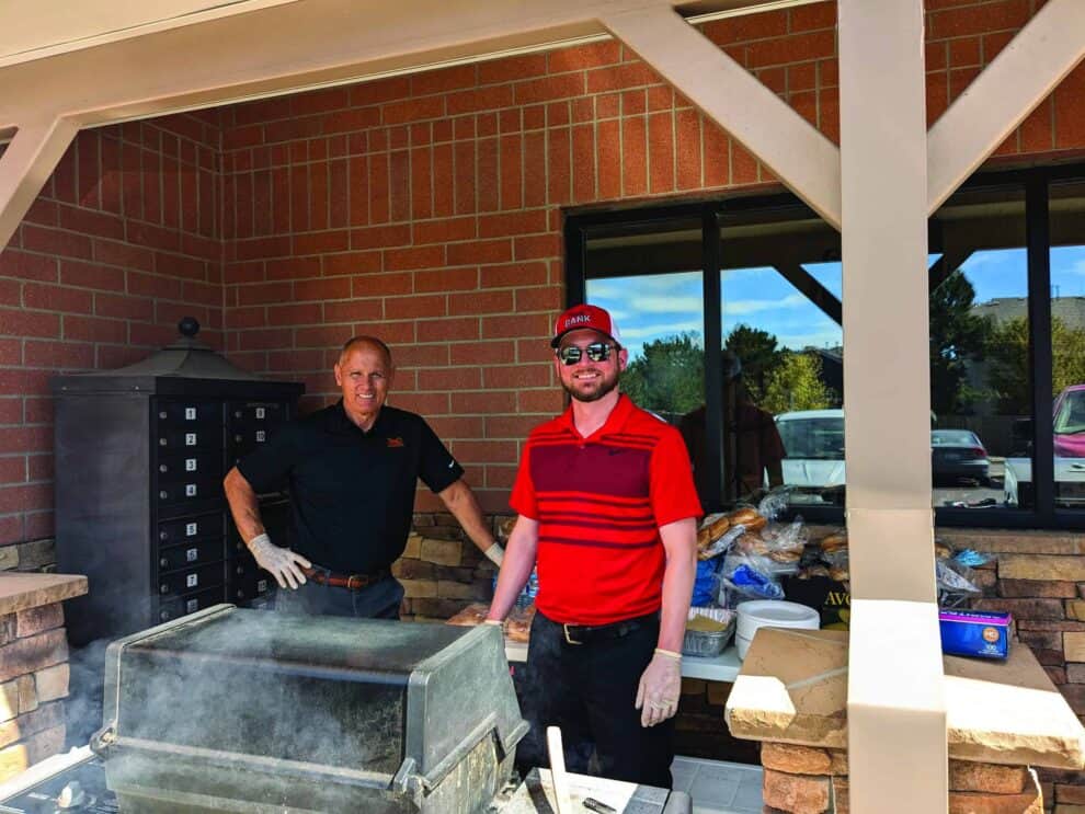 Two people standing by a grill outdoors, one wearing a red shirt and cap, both smiling. Brick building and tables with food items in the background.