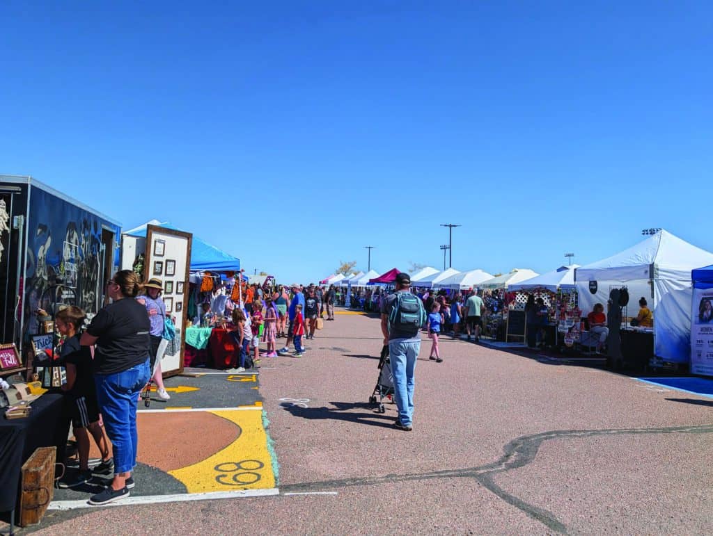 People browse outdoor market stalls under clear skies, with various goods displayed and a central pathway for walking.