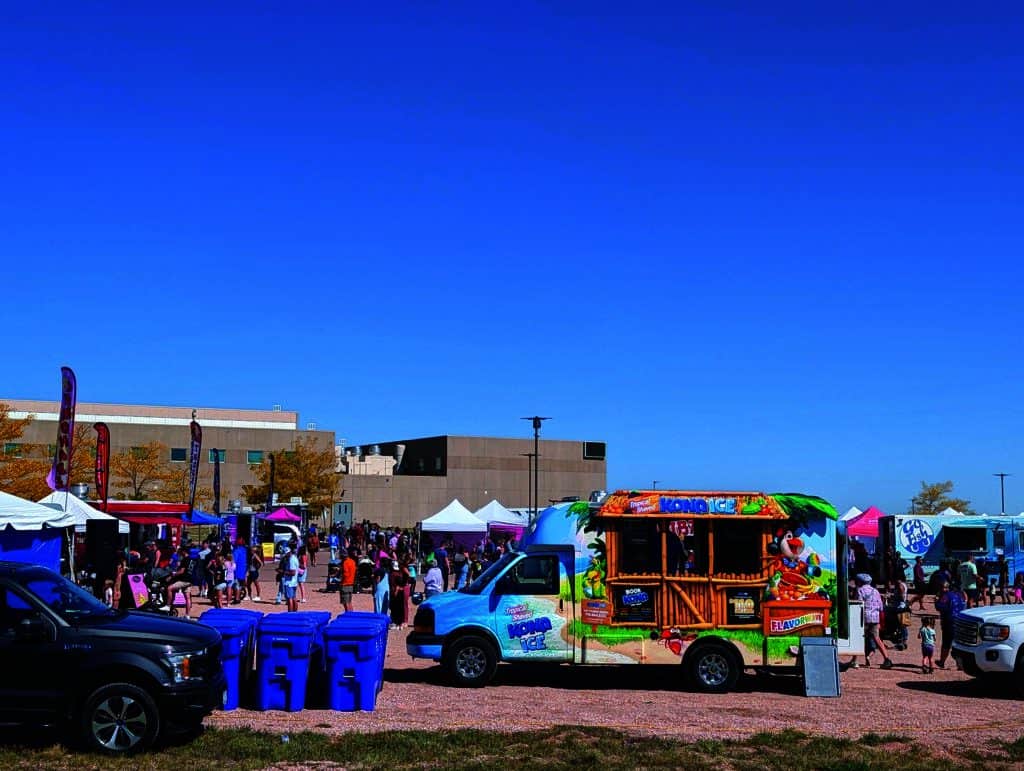 Outdoor festival with food trucks, people in casual attire, blue recycling bins, and tents. Bright, sunny day with clear sky.