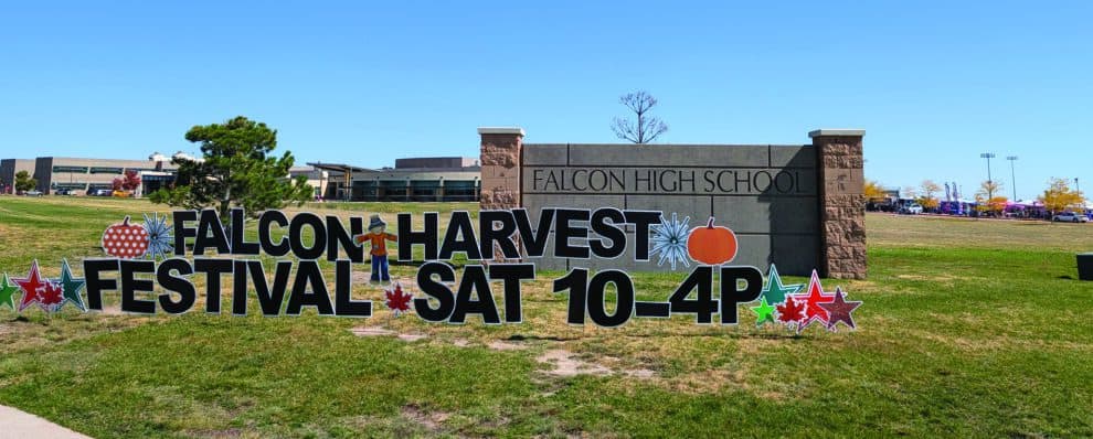 A sign reads "Falcon Harvest Festival Sat 10-4P" in front of Falcon High School, with fall-themed decorations on a grassy area.