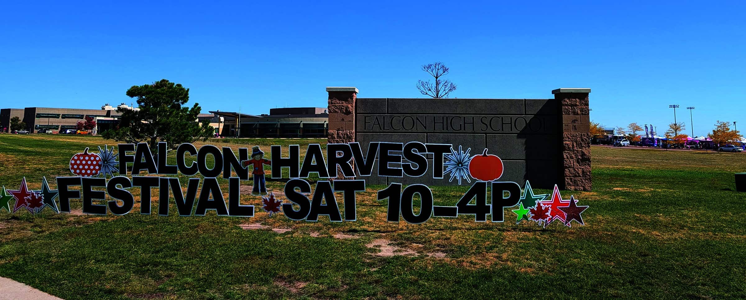 A sign reads "Falcon Harvest Festival Sat 10-4P" in front of Falcon High School, with fall-themed decorations on a grassy area.