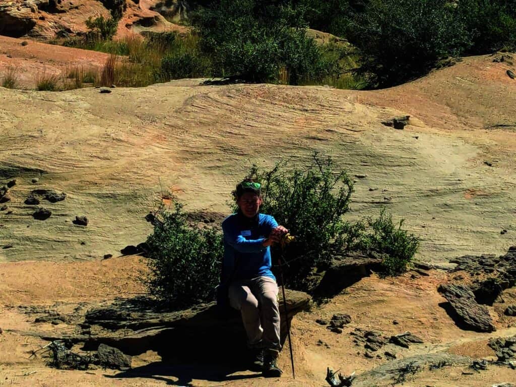 Person sitting on rocky terrain with sparse vegetation, holding a walking stick in a desert landscape.