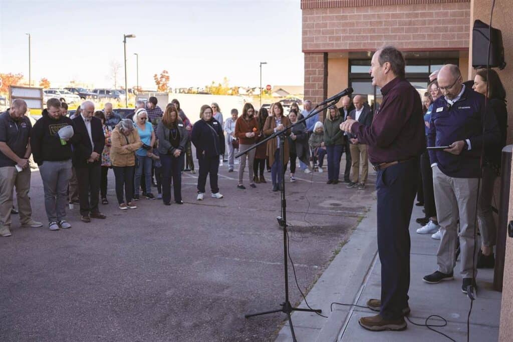 A man speaks into a microphone outside a building while a group of people stands listening.