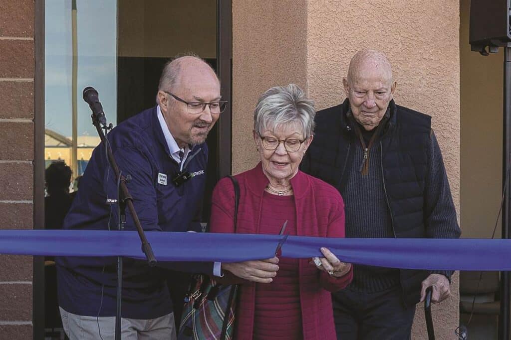 Three people stand in front of a building, where a woman is cutting a blue ribbon with a pair of scissors. A microphone and a brick wall are visible in the background.