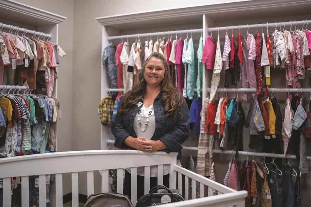 A woman stands smiling in a room filled with neatly arranged children's clothes on racks. A white crib is in the foreground.