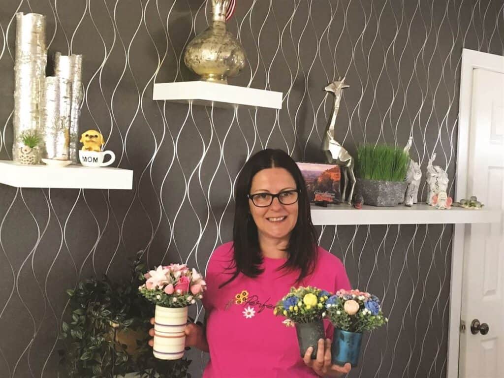 Woman in a pink shirt holds two floral arrangements in small pots, standing in a room with decorative shelves and wall decor.