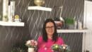 Woman in a pink shirt holds two floral arrangements in small pots, standing in a room with decorative shelves and wall decor.