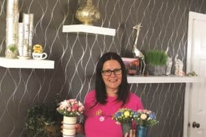 Woman in a pink shirt holds two floral arrangements in small pots, standing in a room with decorative shelves and wall decor.