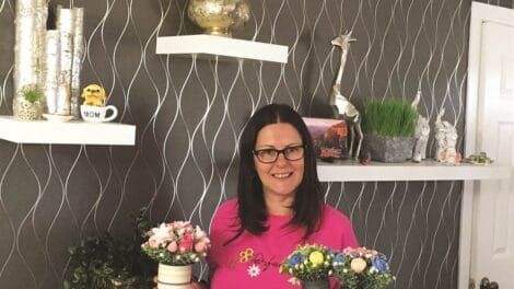 Woman in a pink shirt holds two floral arrangements in small pots, standing in a room with decorative shelves and wall decor.