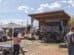 Outdoor music festival with a band performing on a stage. People are sitting and standing under string lights and a large umbrella. Bales of hay are scattered around the area.