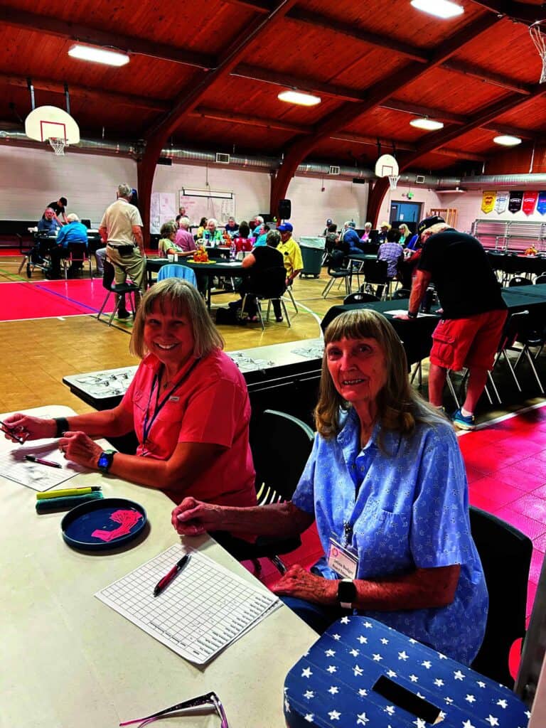 Two women sit at a table with bingo cards in a gymnasium. Several people are gathered at tables in the background, participating in the event.