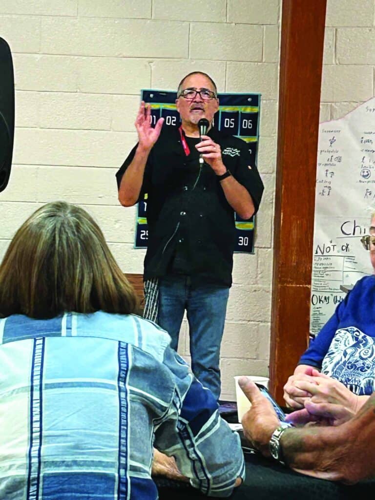 A man speaks into a microphone, gesturing with one hand. Several people are seated facing him in a room with a scoreboard on the wall.