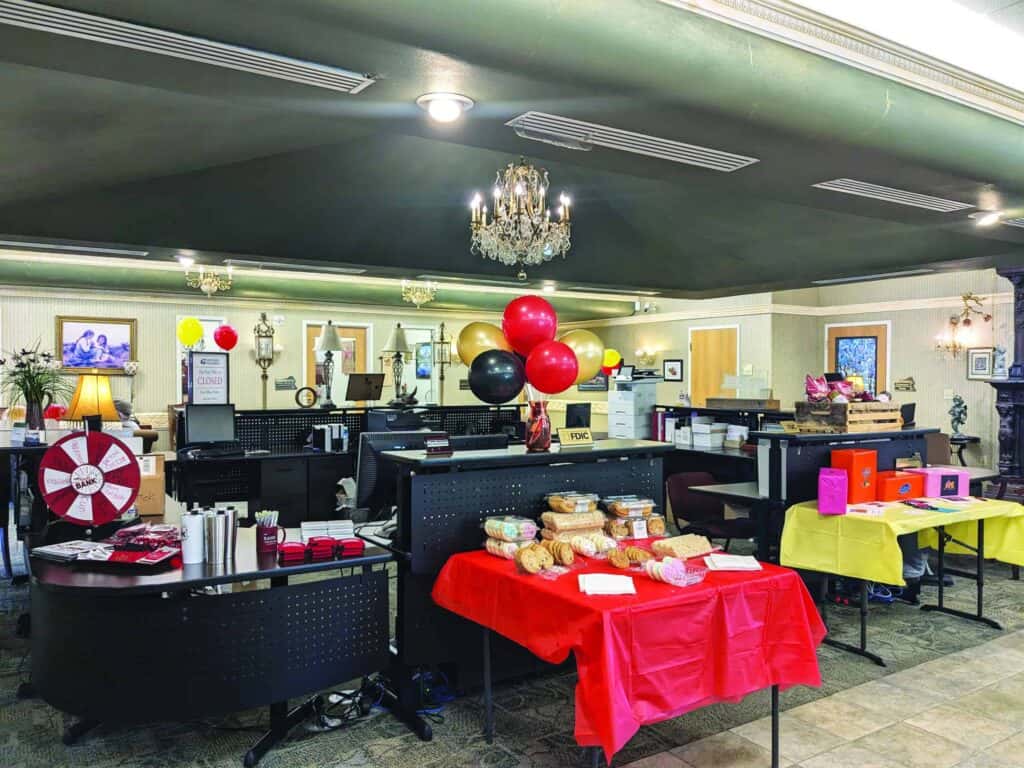 A decorated office space with balloons, tables covered with red and yellow cloths, and various items like food packages and boxes. A chandelier hangs from the ceiling.