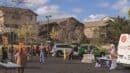 People gather in a parking lot for a community event, with cars and tables set up. Some individuals are wearing costumes, and a person is sitting at a table near a van.