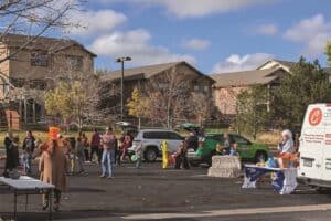 People gather in a parking lot for a community event, with cars and tables set up. Some individuals are wearing costumes, and a person is sitting at a table near a van.