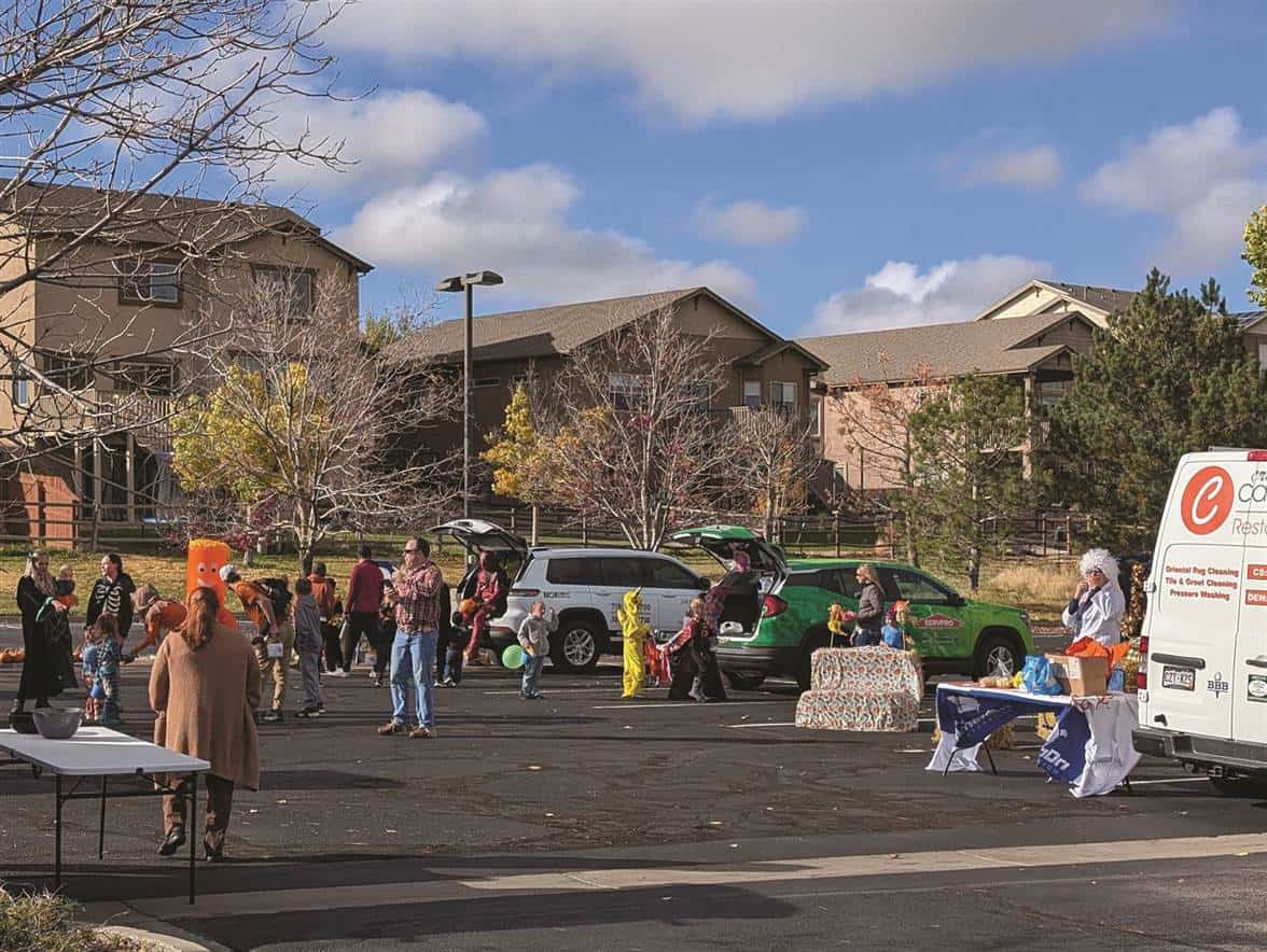 People gather in a parking lot for a community event, with cars and tables set up. Some individuals are wearing costumes, and a person is sitting at a table near a van.