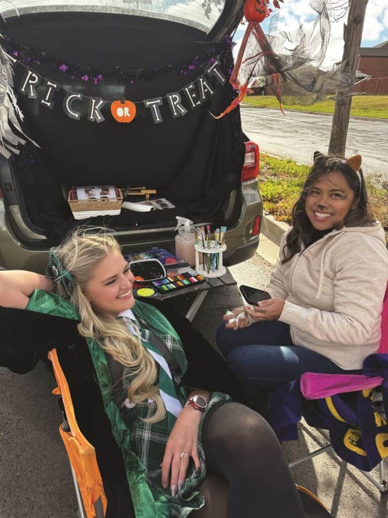 Two women sitting in front of an open car trunk decorated for Halloween with "Trick or Treat" sign. One is in costume; the other wears casual clothes. Art supplies are visible in the trunk.