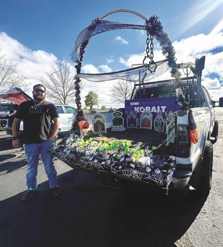 A man stands beside a truck decorated with Halloween-themed items, including tombstones and pumpkins, in a parking lot.