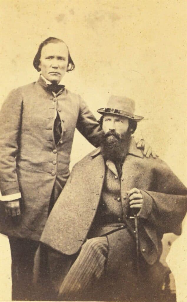 Two men in 19th-century attire, one standing with his hand on the seated man's shoulder. The seated man wears a hat and holds a walking stick. Black and white photograph.