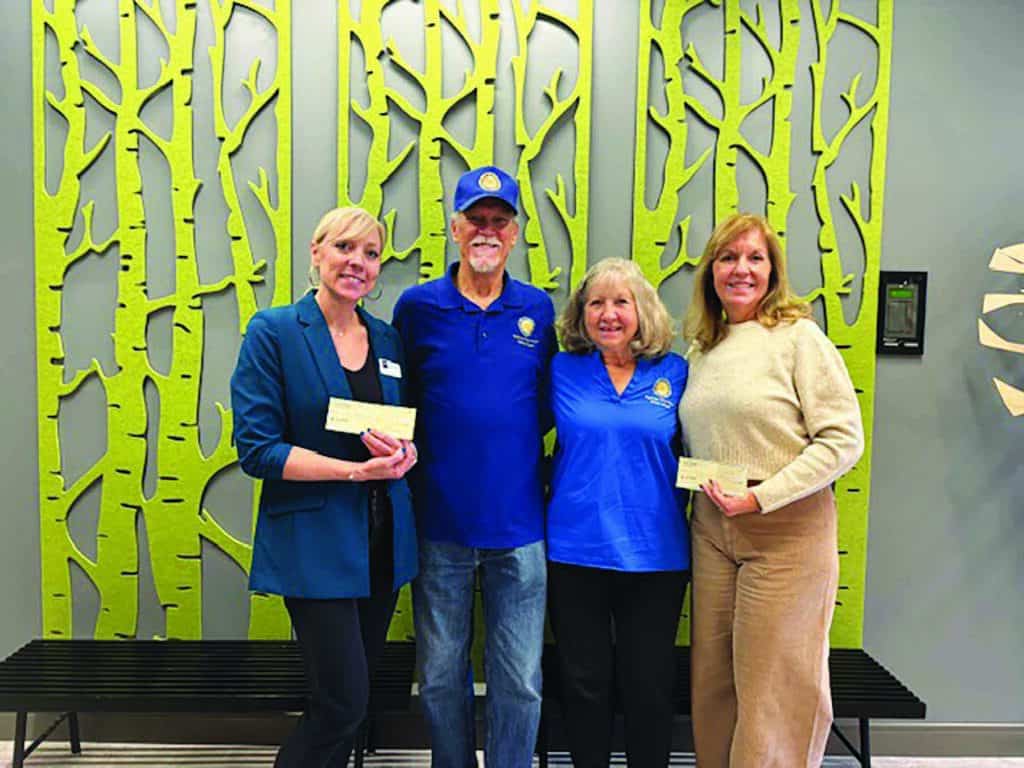 Four people stand smiling in front of a wall with a green tree pattern, holding checks. Two wear blue shirts with a logo.