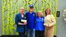 Four people stand smiling in front of a wall with a green tree pattern, holding checks. Two wear blue shirts with a logo.