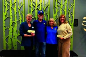 Four people stand smiling in front of a wall with a green tree pattern, holding checks. Two wear blue shirts with a logo.