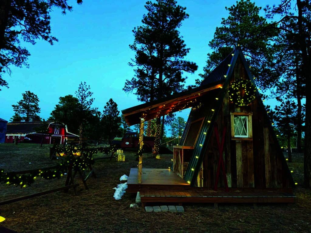 A-frame cabin in a wooded area, decorated with string lights. A barn is visible in the background with surrounding trees and grass.