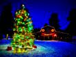 Festive Christmas tree with colorful lights and gifts in a snowy area near a red barn decorated with wreaths and lights, under a deep blue evening sky.