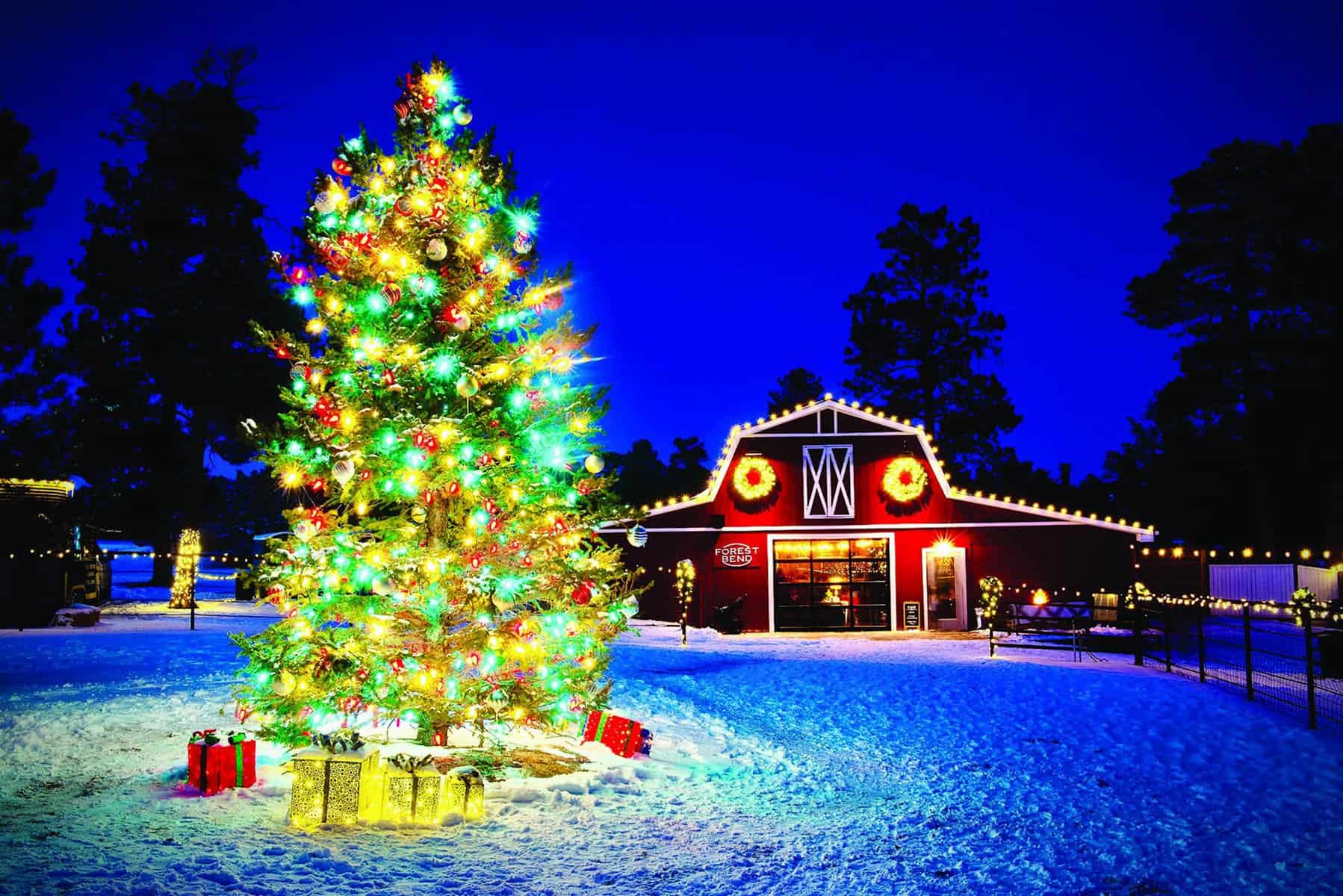 Festive Christmas tree with colorful lights and gifts in a snowy area near a red barn decorated with wreaths and lights, under a deep blue evening sky.
